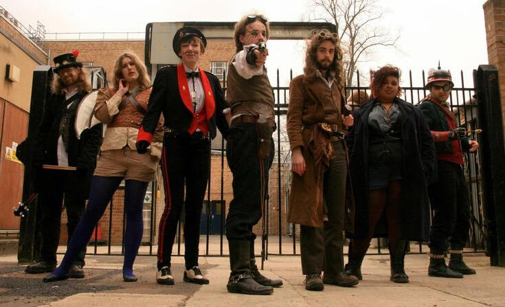 photograph of seven members of the band The Mechanisms. from left to right; Drumbot Brian, Raphaella la Cognizi, The Toy Soldier, Jonny d'Ville, Gunpowder Tim, Ashes O'Reilly, Marius von Raum. Jonny is pointing a gun at the camera and there's a fence behin